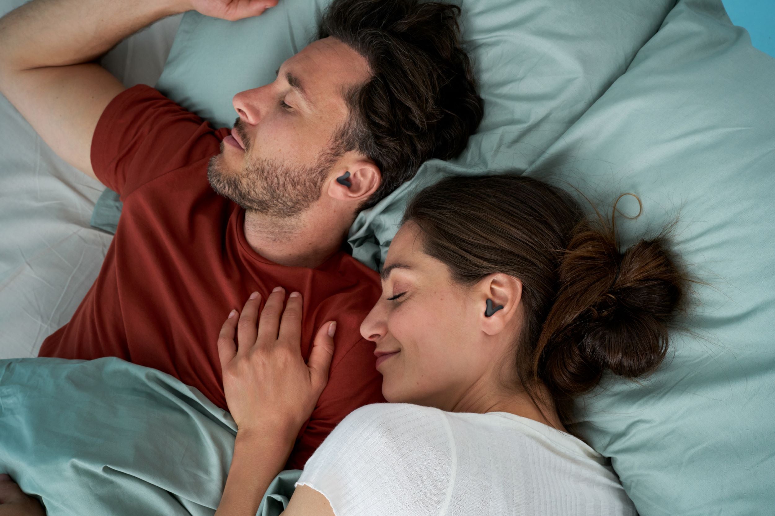 Young couple laying on the bed next to each other, both of them with Alpine Silence Black in their ears and smiling. The woman has her hand on the man& chest