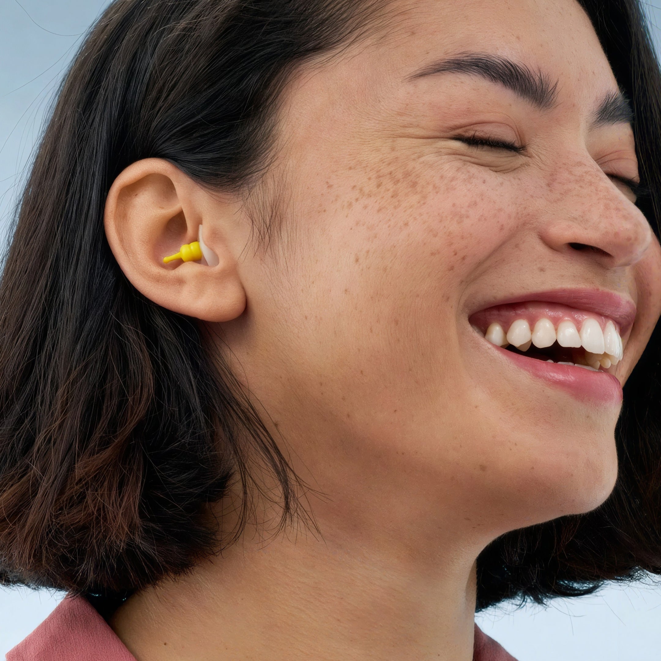 Alpine FlyFit earplug worn in the ear by a smiling woman with dark shoulder-length hair 